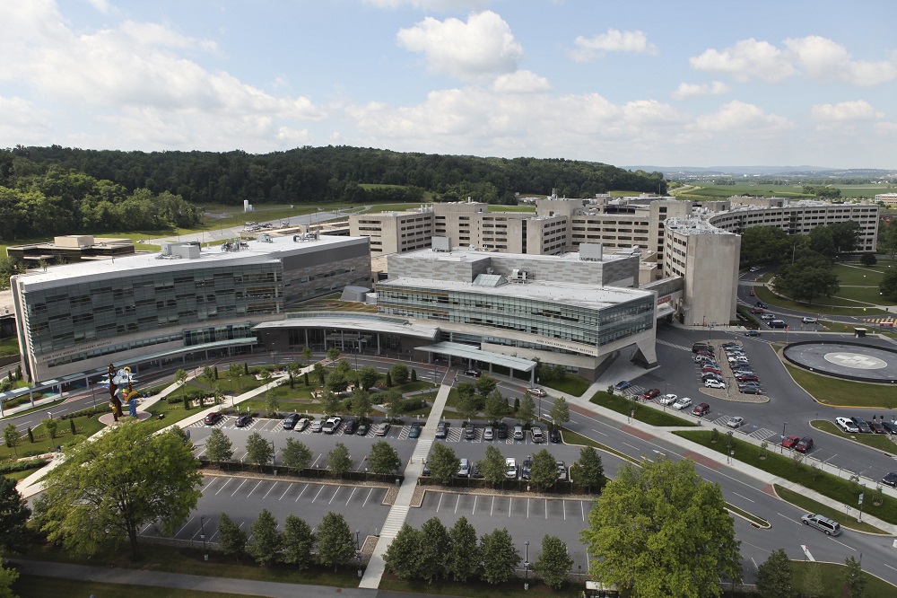 An aerial view of Penn State Health Milton S. Hershey Medical Center and Penn State College of Medicine shows tall buildings, a parking lot with cars in the foreground and hills with trees in the background.