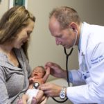 A physician from Penn State Health Medical Group ― Kissell Hill listens to a newborn baby’s heartbeat with a stethoscope. The doctor is wearing a lab coat with “Kissell Hill Family Care” on the sleeve. A woman with long hair is holding the baby and smiling at it. She is wearing a sweater.
