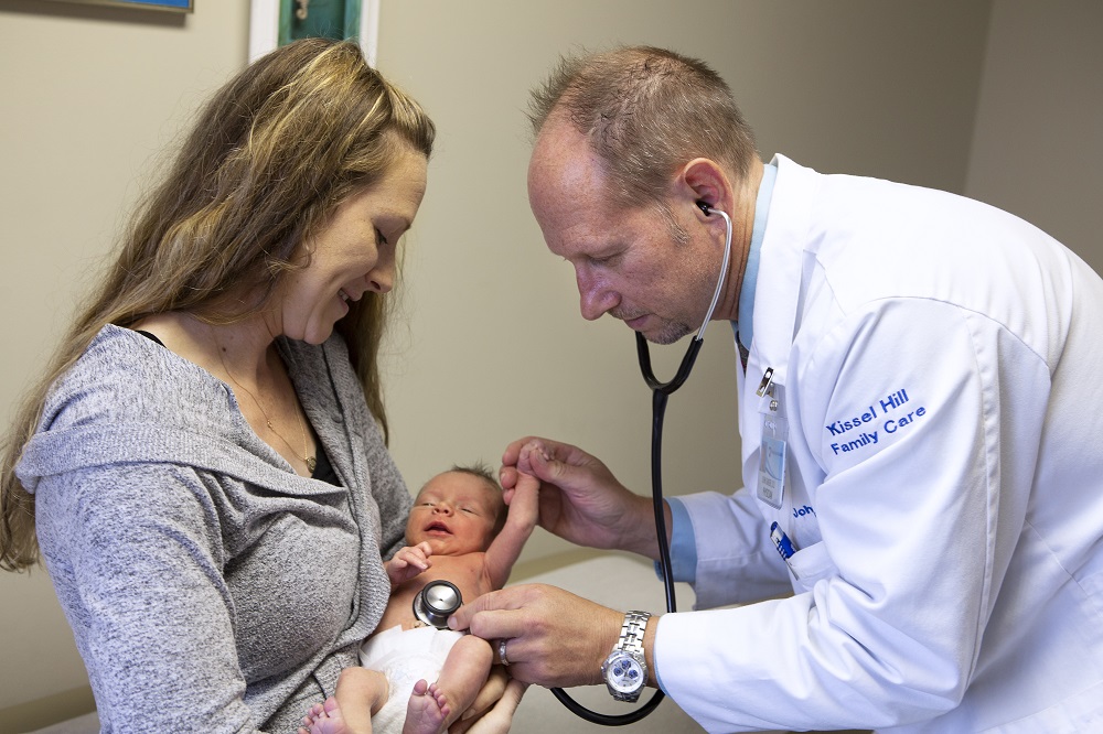 A physician from Penn State Health Medical Group ― Kissell Hill listens to a newborn baby’s heartbeat with a stethoscope. The doctor is wearing a lab coat with “Kissell Hill Family Care” on the sleeve. A woman with long hair is holding the baby and smiling at it. She is wearing a sweater.