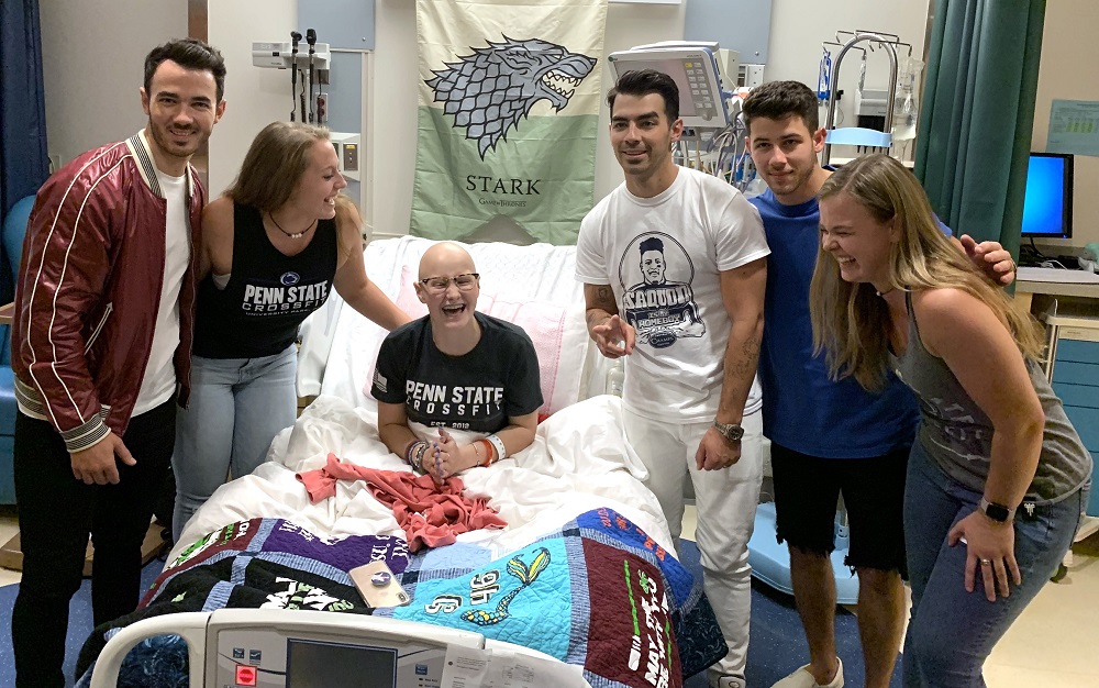 Wearing a Penn State CrossFit shirt and sitting up in a hospital bed, Lily Jordan laughs. Next to her are two women and three young men, the Jonas Brothers.