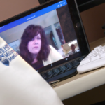 A close-up of Cynthia Lacey’s face is on a laptop monitor that is angled to the left. Dr. Simmons arm with the Hershey Medical Center logo on it is visible in the foreground. Next to him is a keyboard.