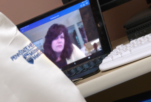 A close-up of Cynthia Lacey’s face is on a laptop monitor that is angled to the left. Dr. Simmons arm with the Hershey Medical Center logo on it is visible in the foreground. Next to him is a keyboard.