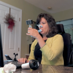 ALS patient Cynthia Lacey holds a spirometer in her mouth. Noseplugs are on her nose. She is looking into a laptop computer. Next to her a man is standing with his hands crossed. Behind her a living room is visible.