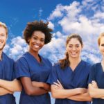 One male nurse and three female nurses stand in a row with their arms crossed. They are wearing blue surgical smocks and smiling.