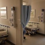 Two hospital beds sit in a room separated by a partition wall. Curtains hang on portions of the wall.