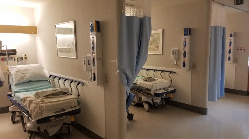 Two hospital beds sit in a room separated by a partition wall. Curtains hang on portions of the wall.