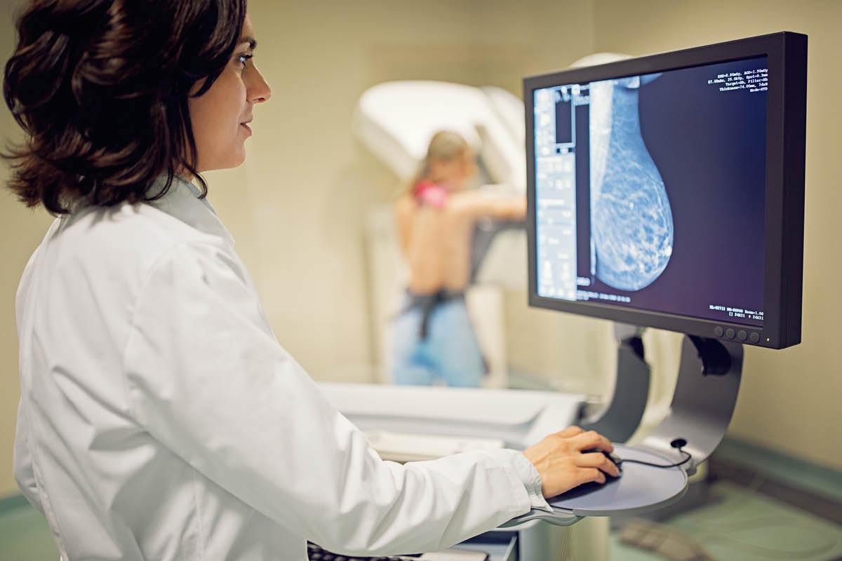 A woman in a medical coat stands at a computer. A mammogram image is on the monitor. In the background, out of focus, a woman stands facing imaging equipment.