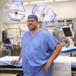 Glenn Weidler leans against an operating room table. He is wearing scrubs and a pair of glasses.