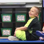 Shane sits cross-legged on a floor. He smiles. The carpet is printed with letters. Next to him are bins labeled Bears, Forest Animals and Zoo Animals.