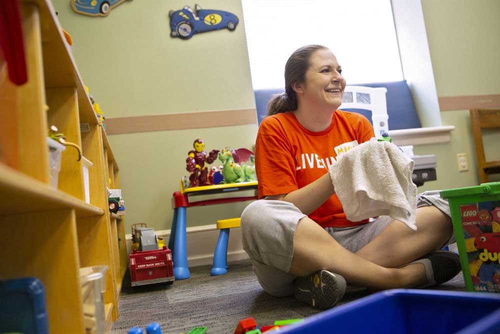 Mary Lucas, wearing a “Live United” T-shirt, sits cross-legged on a carpeted floor. She smiles as she cradles a toy in a washrag. Toys are stacked on shelves and tables around her.