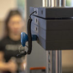 The weights of a weight machine are seen in a close-up image, with a person using the machine out of focus in the background.