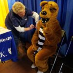 Someone wearing Penn State Nittany Lion costume points holds up a No. 1 sign as Penn State Health nurse Renee Houser pretends to give an injection into the person’s arm. A table draped with a Penn State Health table cloth is next to Houser.