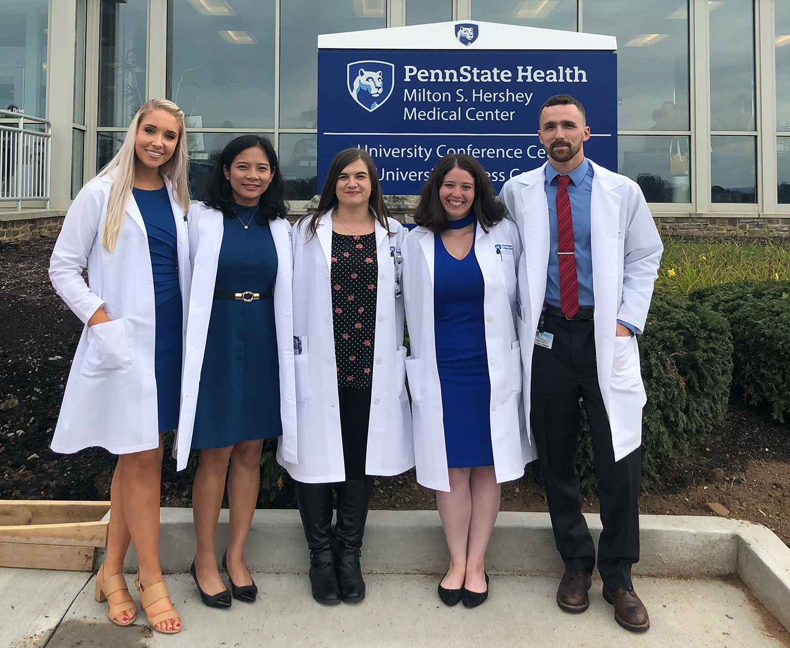 Five graduate students are seen wearing long laboratory coats and dress clothes, standing in front of a Penn State Health conference center in August 2019.
