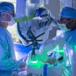 Two people in surgical scrubs look into a robot while performing a surgery