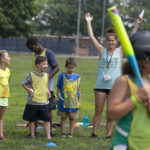 A young girl stands in the foreground holding a baseball bat and wearing a helmet, smiling. In the background, four children and one adult stand, smiling and cheering her on.