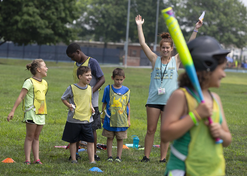A young girl stands in the foreground holding a baseball bat and wearing a helmet, smiling. In the background, four children and one adult stand, smiling and cheering her on.