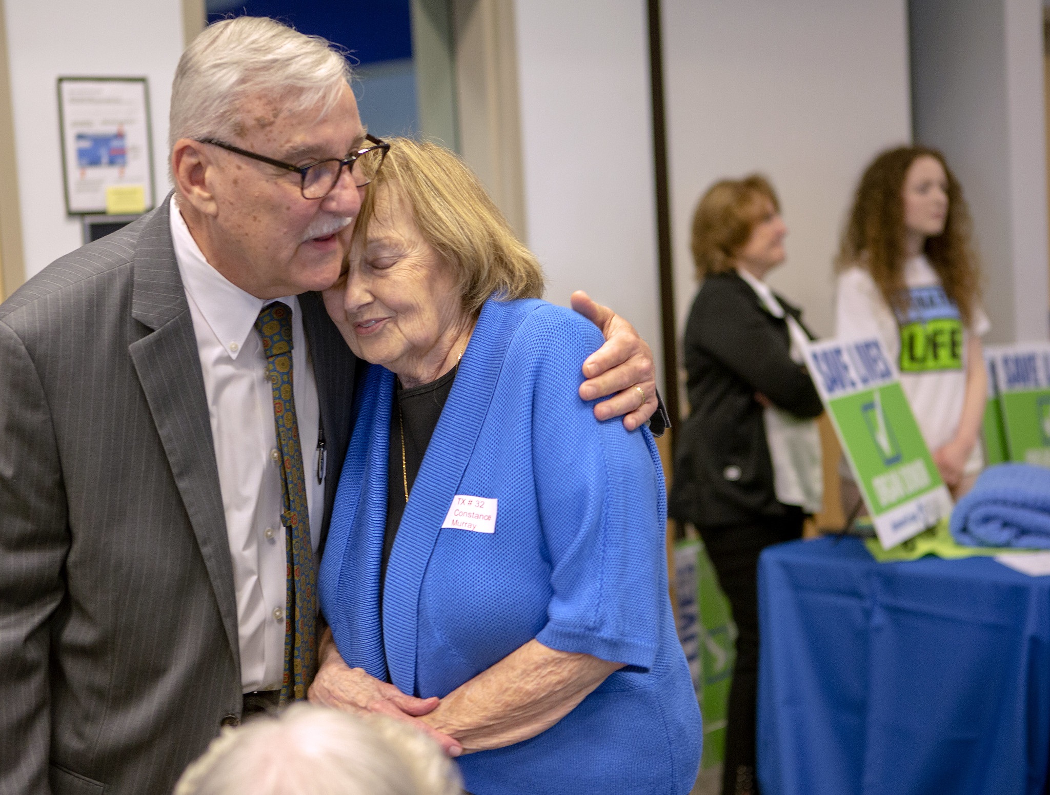Dr. John Pennock, wearing a sport jacket and tie, wraps an arm around Constance Murray. Both are standing in a room surrounded by other people with signs and T-shirts that say “Save Lives.”