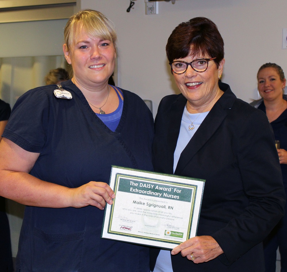 Maike Sgrignuoli, wearing scrubs, and Judy Himes, wearing a suit and glasses, smile as they each hold a corner of a plaque with the words “The DAISY Award For Extraordinary Nurses.”