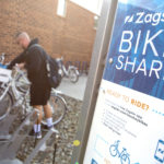 A man reaches for a bike that is in a bike rack. He is wearing a sweatshirt, shorts and a backpack. In the foreground is a sign that says “Zagster Bike Share. Ready to ride?”