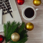 An overhead close up of a wooden desk or tabletop is shown. An open laptop computer sits on the surface, and on the keyboard someone has placed a notepad and a pen. A cup of coffee sits next to keyboard. Also on the surface are pine cutting and holiday decorations.