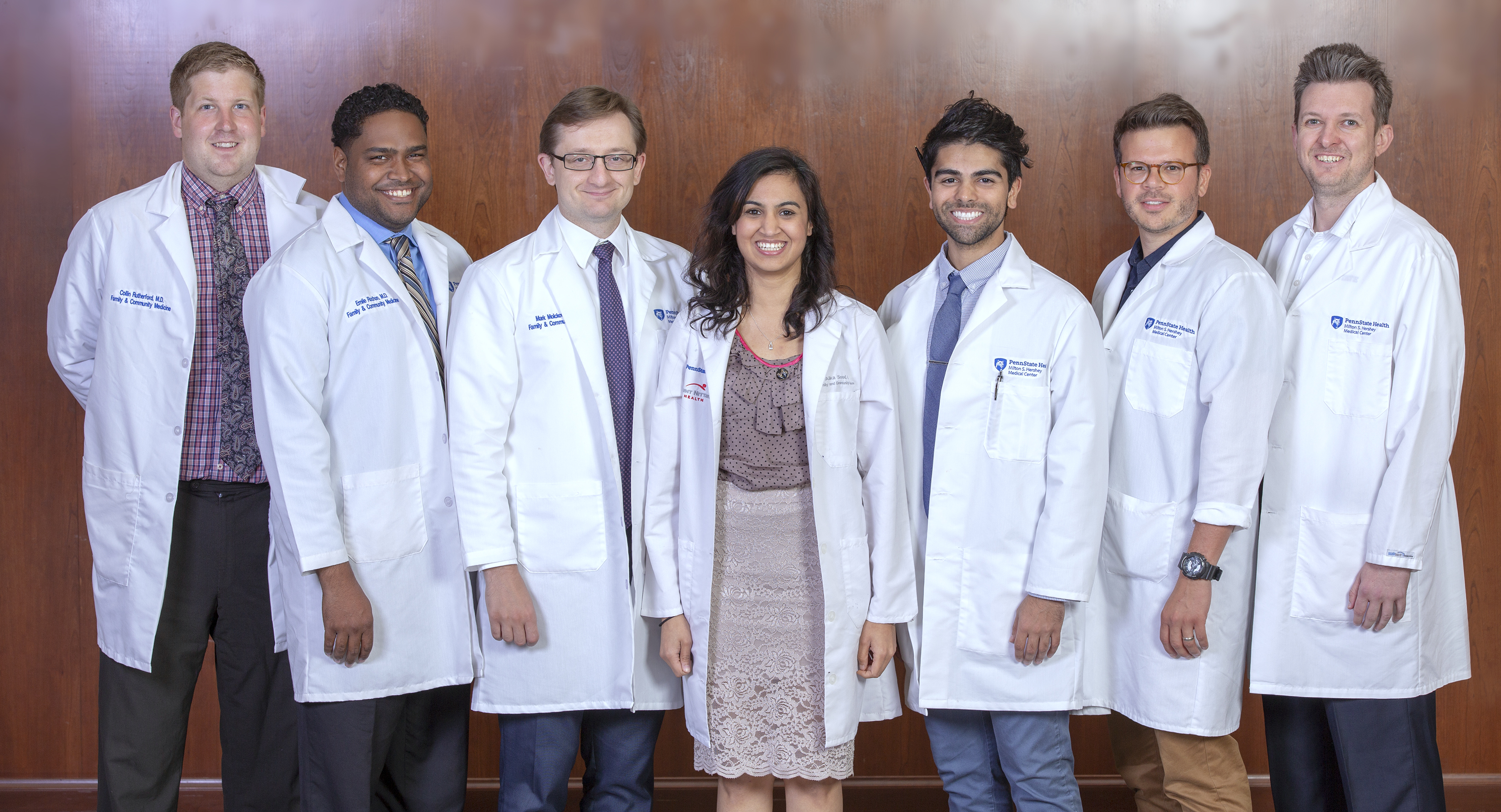 Six men and one woman wearing white lab coats with the Hershey Medical Center logo and their names on them stand in a row and smile. The men are wearing shirts and ties, and the woman is wearing a skirt and top. The wall behind them is made of wood paneling.