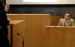 Dr. Mark Molckovsky sits in a witness stand and listens to a woman talking to him. He is wearing a suit and glasses. The woman is seen in profile and she is learning on a podium. A man with a robe, shirt and tie stands to the left of the judge’s seat.