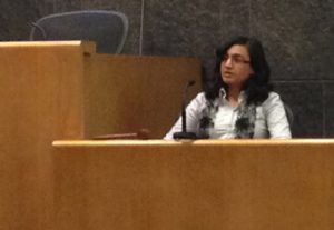 Dr. Alka Sood sits on a witness stands and speaks into a microphone. The witness stand is made of wood and is lower than the judge’s seat to her left. She has shoulder-length hair and is wearing glasses, a blouse and a scarf.