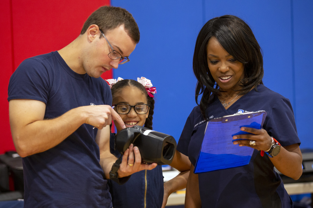A man holds a piece of vision screening equipment, pointing to it, as a young girl looks on. A woman stands on the other side of the girl, holding and looking at a clipboard.