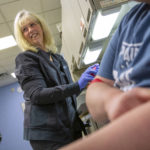 Cindy Twombley stands and smiles as she looks to the left. She has long hair and is wearing a sweatshirt and rubber gloves. Next to her a female patient wearing a T-shirt sits with an armband around her arm.