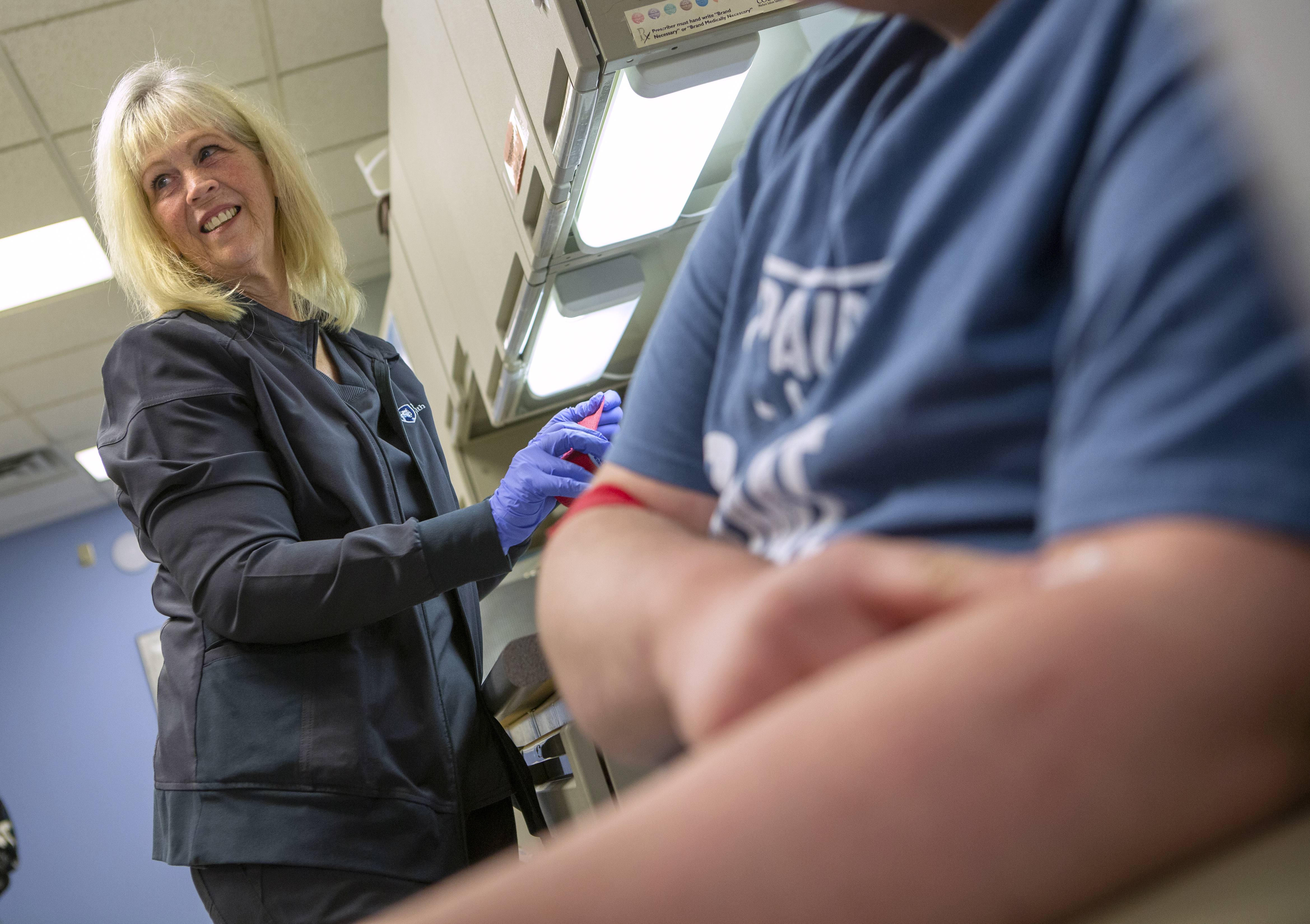 Cindy Twombley stands and smiles as she looks to the left. She has long hair and is wearing a sweatshirt and rubber gloves. Next to her a female patient wearing a T-shirt sits with an armband around her arm.