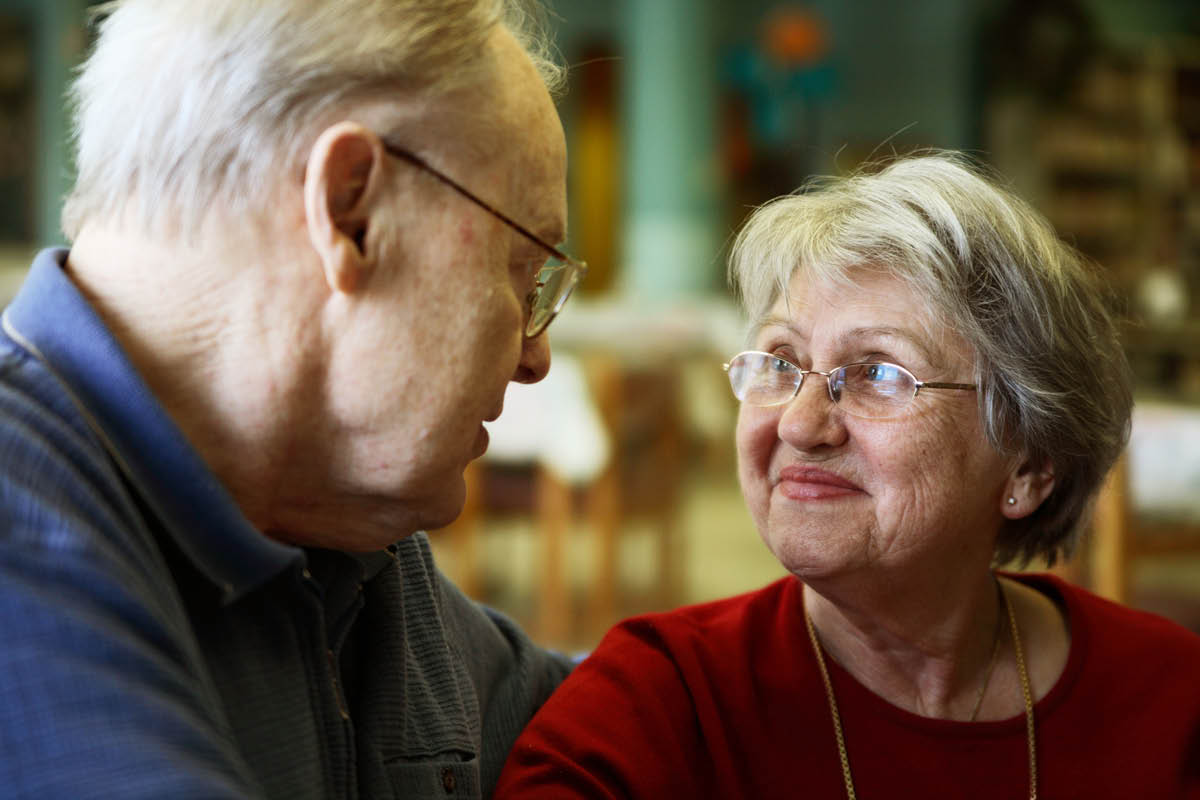 A man and a woman sit next to each other. The man has his arm around the woman. The two look at each other, smiling slightly.