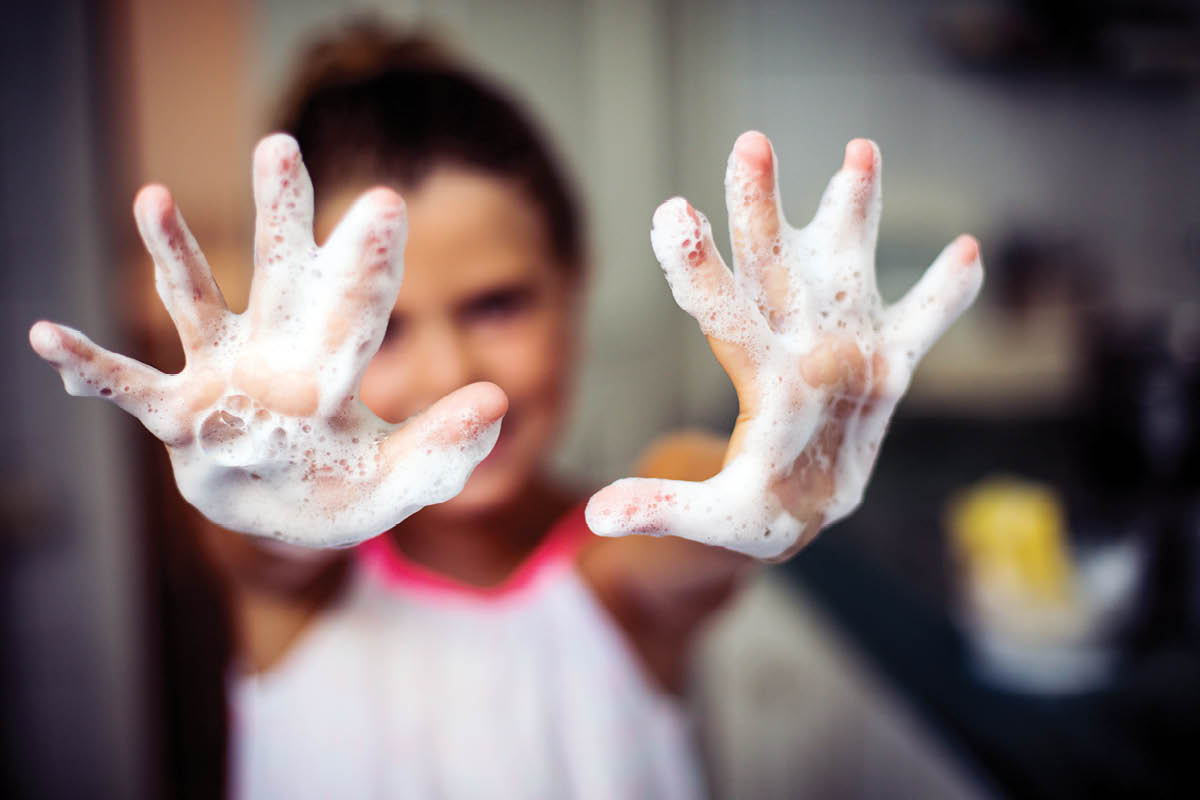 A young child stands with both hands stretched directly toward the camera. Both hands are covered in soap suds. The rest of the child, in the background, is blurry.