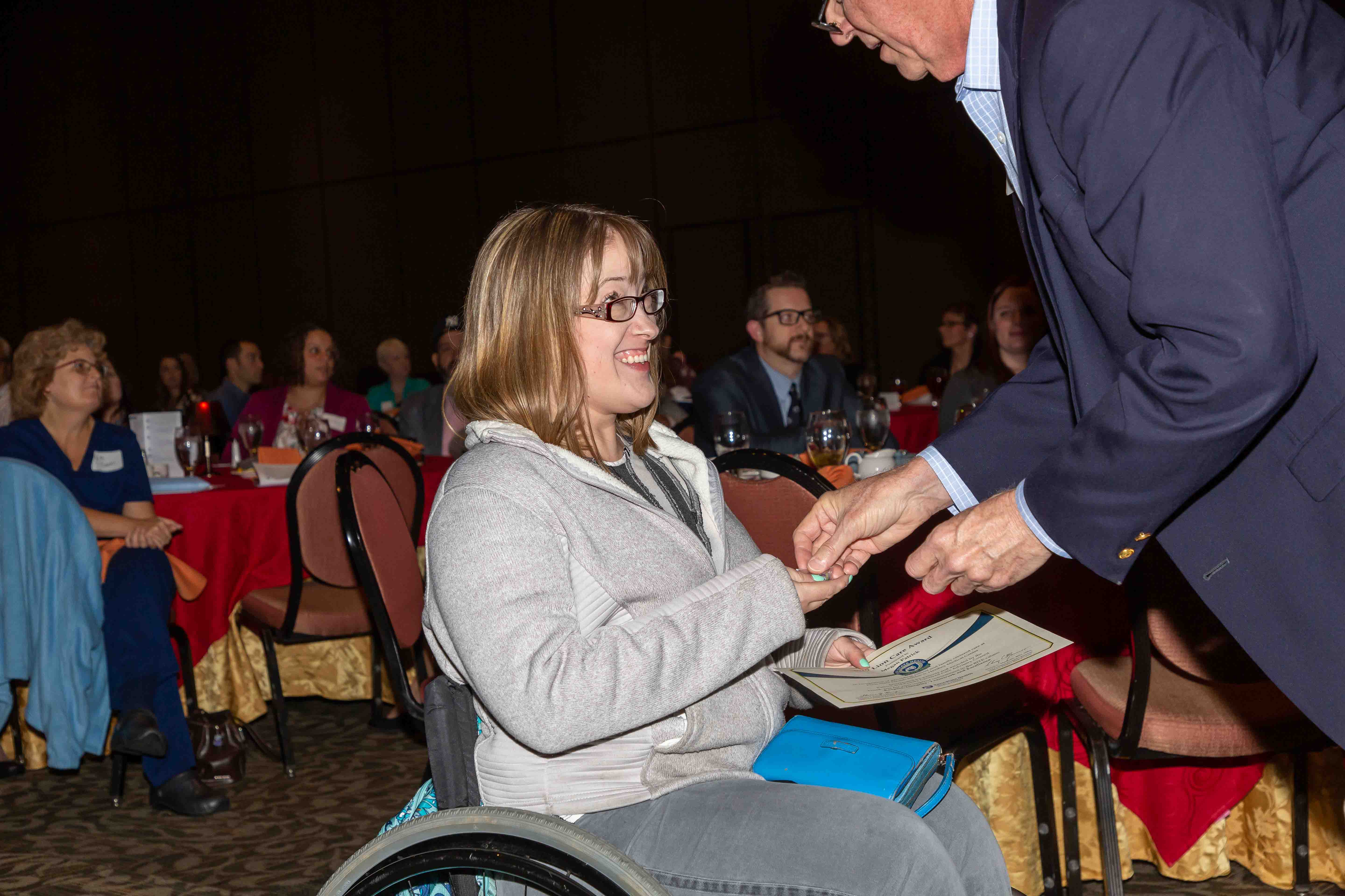 Megan Patrick, wearing a sweatshirt and jeans, smiles at a man in a sport coat who is placing something in her hand. In her other hand, she holds a certificate.