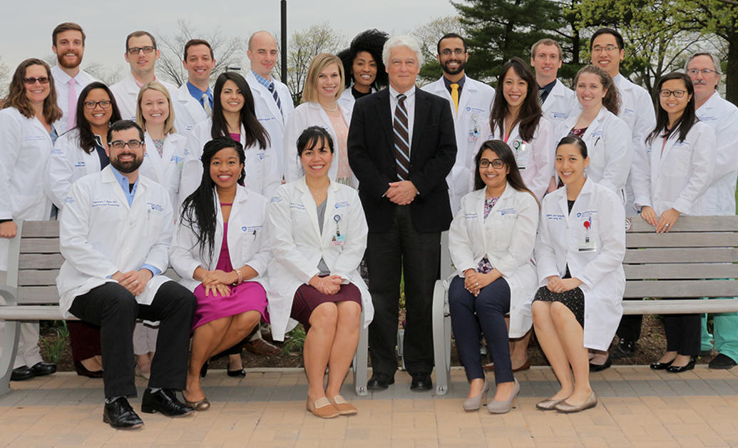 Dr. John Repke, wearing a suit, poses outdoors with 26 residents - some seated, some standing -- all of whom wear white clinical coats.