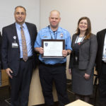 Five people stand in a row. A man in the center wearing a police-style uniform holds a plaque.