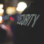 A close up of someone with word security on his shirt in profile stands against lights out of focus