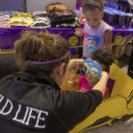 Child Life Assistant Tiffaney Horner places special glasses on the face of 2-year-old Ava Lawson as her sister Aurora, 3, looks on. Ava is seated in a toy car. In the near background is a table with sunglasses, shirts, cups and other merchandise on it.