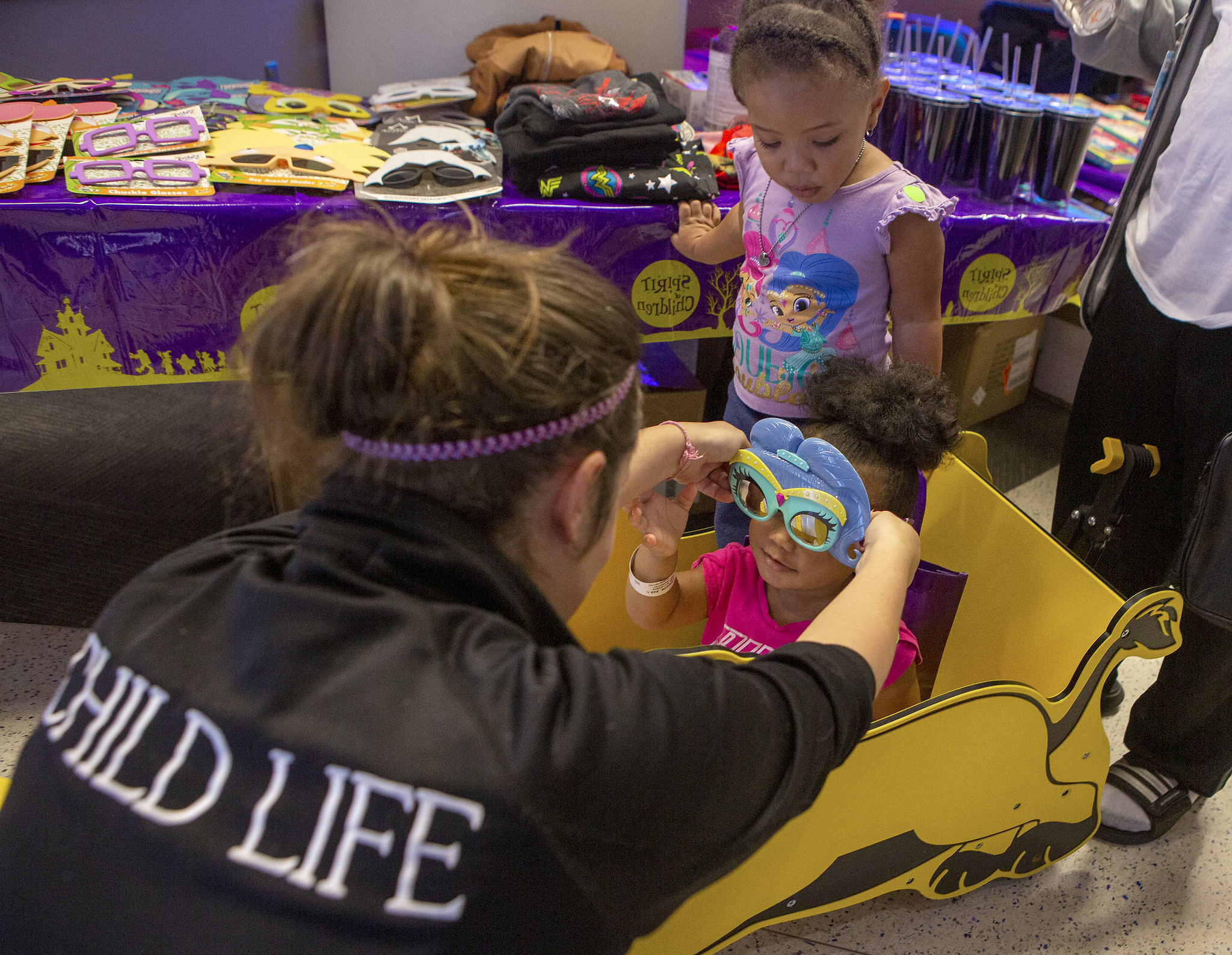Child Life Assistant Tiffaney Horner places special glasses on the face of 2-year-old Ava Lawson as her sister Aurora, 3, looks on. Ava is seated in a toy car. In the near background is a table with sunglasses, shirts, cups and other merchandise on it.
