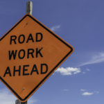 A diamond-shaped roadwork sign stands against a blue sky.