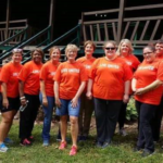 A row of men and women stand in front of a building with porch with a wood railing. All the men and women are wearing “Live United” T-shirts.