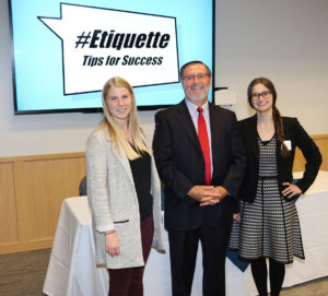 Three people are seen standing in front of a table. A digital presentation screen behind them displays the hashtag Etiquette.