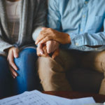 A man and a woman sit side-by-side on a couch. Image shows them from the neck down. The woman’s left hand is on the man’s right leg, which he holds with both hands.