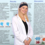 A medical student wearing a medical coat is pictured with an out-of-focus scholarly poster of hers visible in the background.