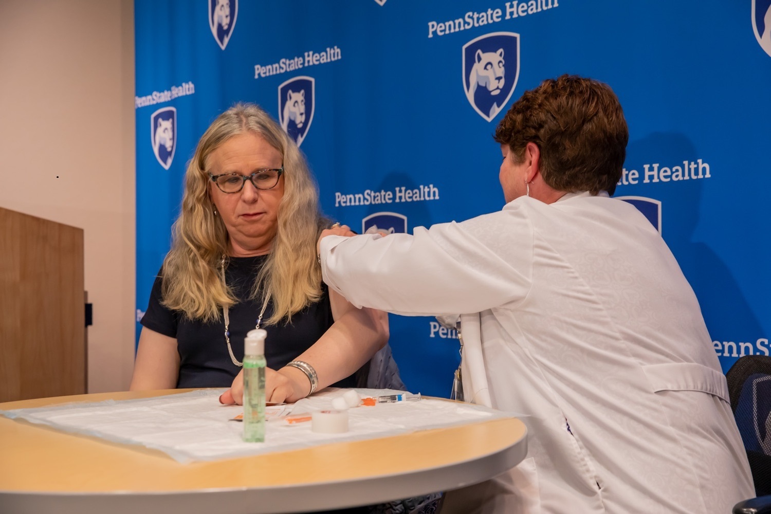 Dr. Rachel Levine, left, sits at a table with Lori Bechtel, nurse manager for Employee Health and Employee Safety as Bechtel gives her a flu shot. Levine has long, wavy hair and is wearing glasses and a dress. Bechtel has short hair and is wearing a white lab coat. Hand sanitizer and a syringe are on the table. Behind them is a banner with the Penn State Health logo on it and a podium.