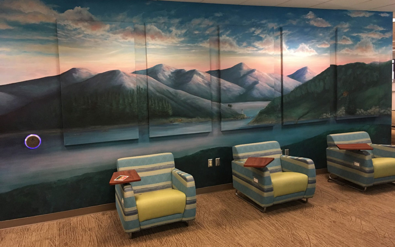 A multi-panel mural depicting mountains with an ocean in front of them and a sky above them is seen in a room with large, comfortable chairs in front of it.
