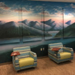 A multi-panel mural depicting mountains with an ocean in front of them and a sky above them is seen in a room with large, comfortable chairs in front of it.