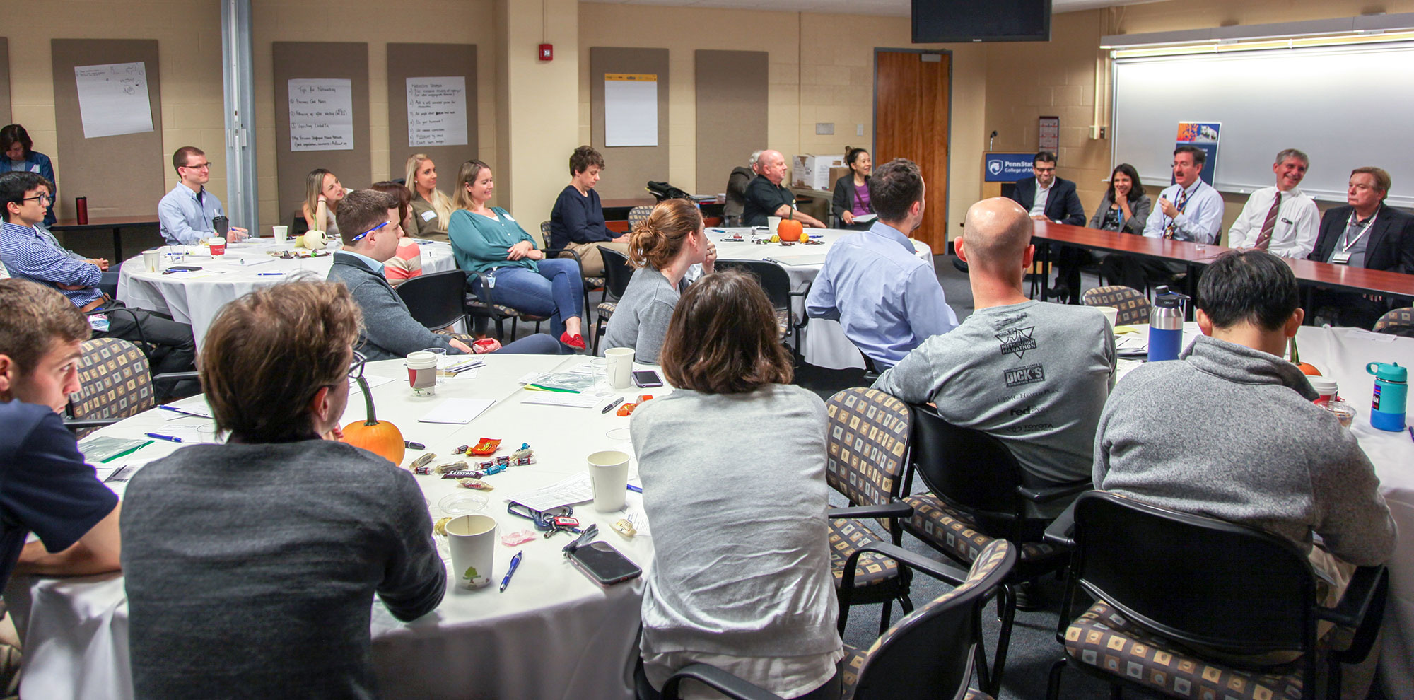 A group of people are seen sitting at round tables in a large room. A panel of speakers sits across a table at the front of the room.