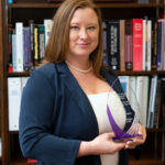A professional photo of Jennifer Nyland, PhD, shows her holding an award as a Woman to Watch in STEM. She is standing in her office with books and a framed degree in the background.