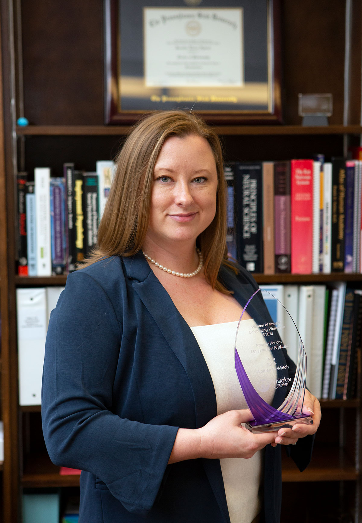 A professional photo of Jennifer Nyland, PhD, shows her holding an award as a Woman to Watch in STEM. She is standing in her office with books and a framed degree in the background.
