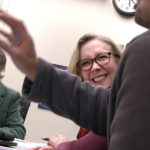Physician Writers Group founder Kimberly Myers and Dr. Eileen Moser look at Dr. Jose Stoute as he presents an original work during a workshop in February. They are seated at a table with papers and laptops on it. Myers has short brown hair and glasses and is wearing a blazer with a turtleneck shirt and a scarf tied loosely around her neck. Moser, who wears glasses, smiles broadly at Stoute, who is gesturing with his hand. His face is not shown. A clock is on the wall behind them.